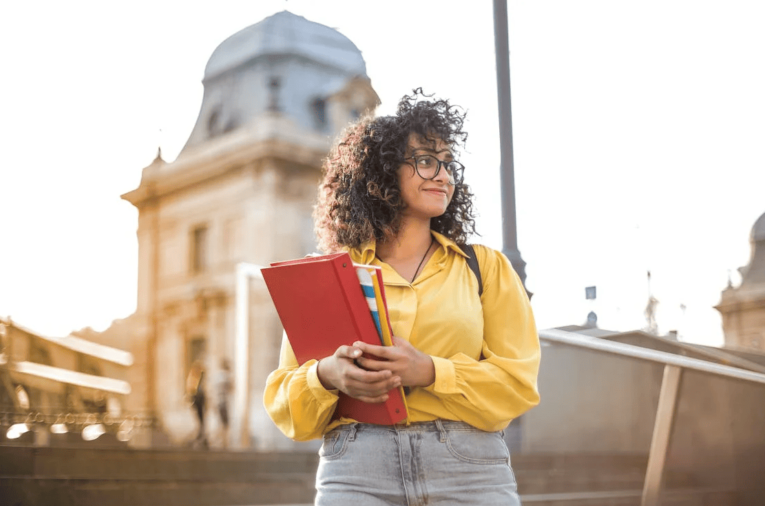 A student with a yellow jacket holding books and standing in front of a building.