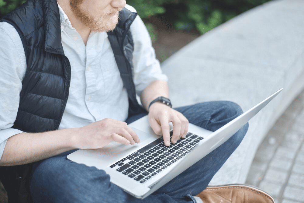 A person typing on their laptop while sitting on a concrete bench outdoors.