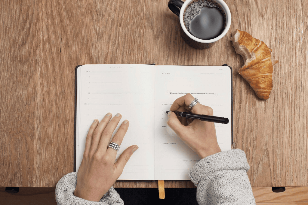 A person writing in a notebook with a cup of coffee and a half-eaten croissant next to them.