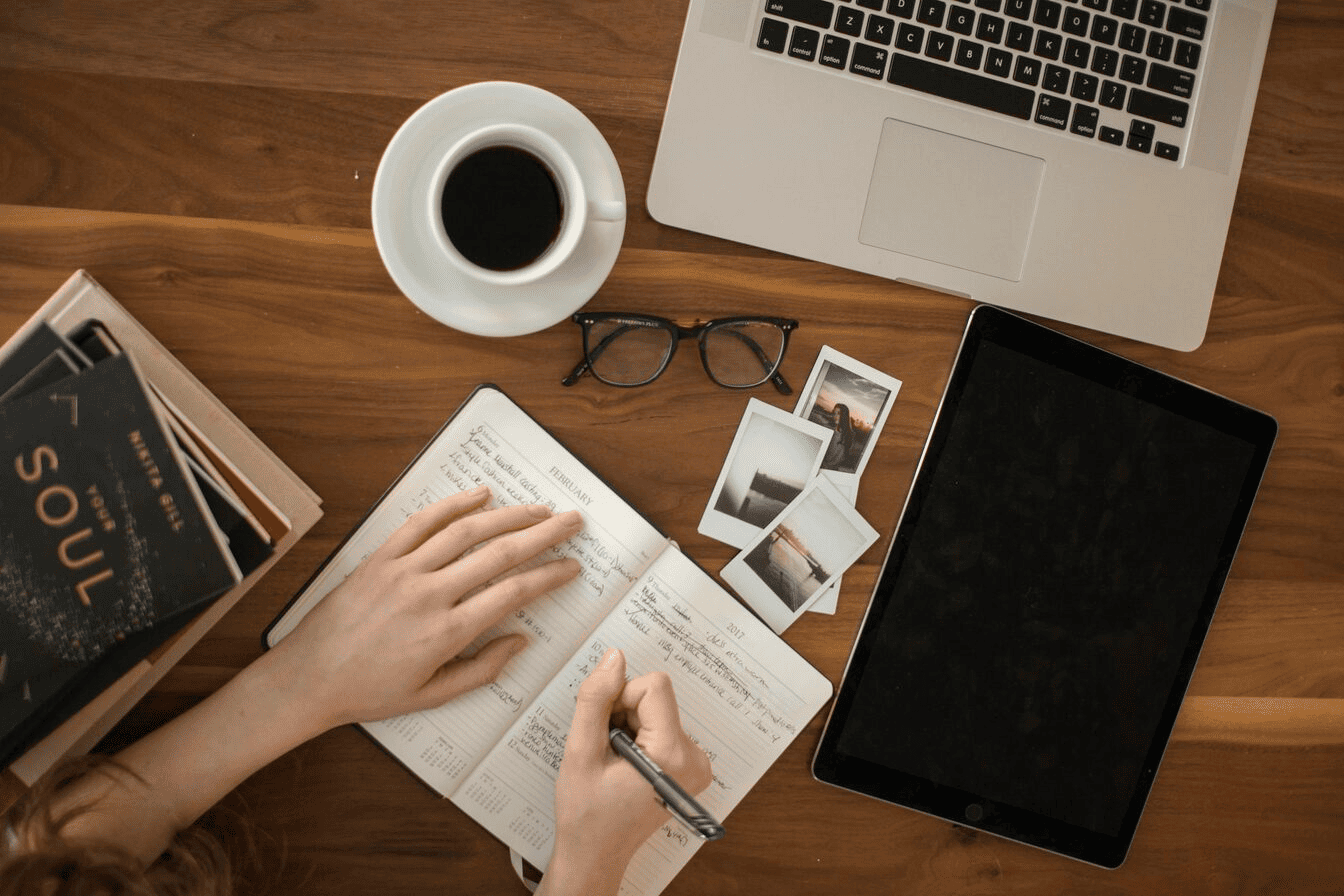 A person holding a ballpoint pen and writing on a notebook with a laptop, tablet, and coffee mug next to them.