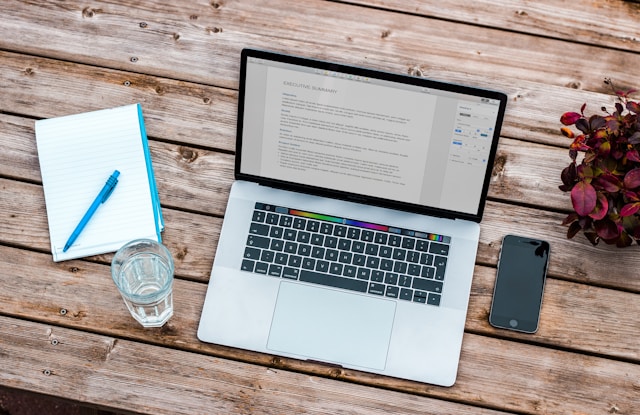 A laptop, a smartphone, a glass of water, a notebook, and a plant on a wooden table. 