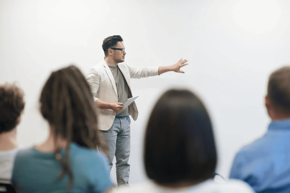 A teacher in front of students pointing at a white wall.