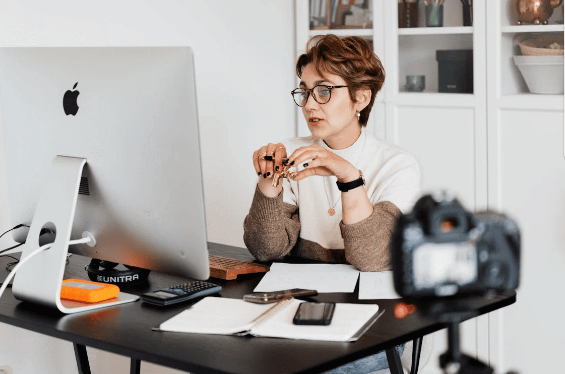 A woman looking at her Mac computer monitor with a camera opposite her.