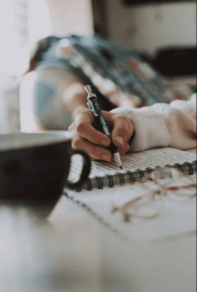 A person writing in a notebook with cup of coffee next to them.