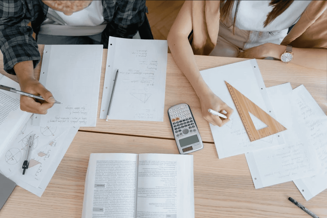 Students working on a project while writing notes with pieces of paper, a book, and a calculator next to them.