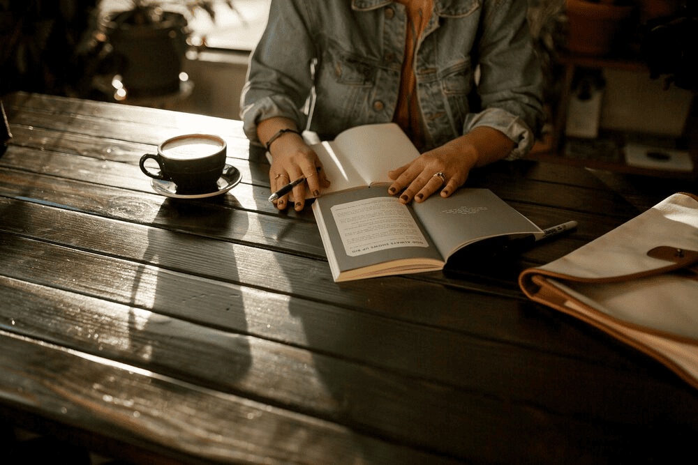 A person looking at a book and making notes while working on a brown wooden table.