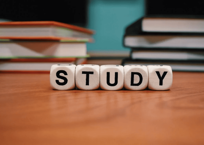 Five dice spelling the word "study" on a wooden desk in front of some books.