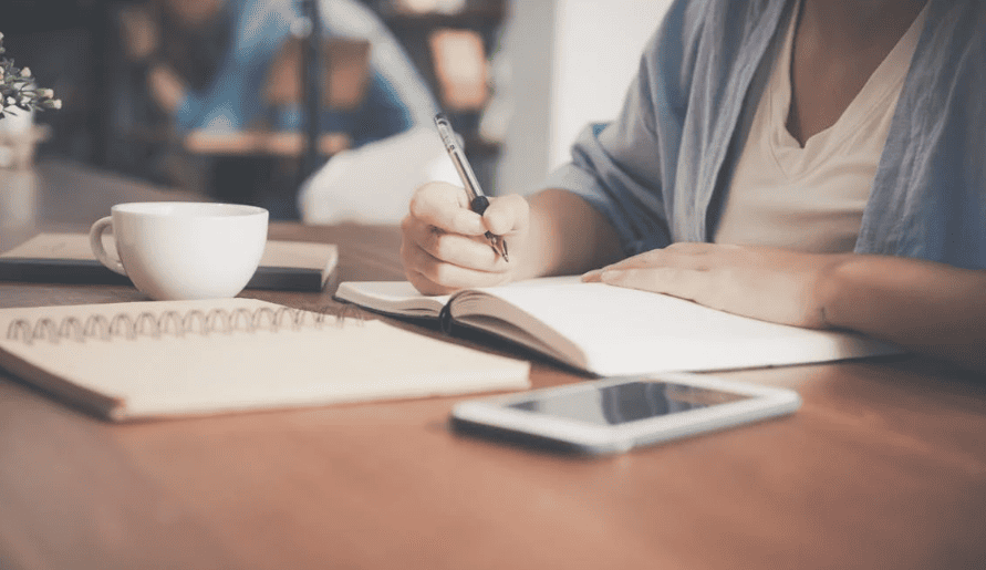 A woman writing in a notebook with a cup of coffee and a smartphone next to her.