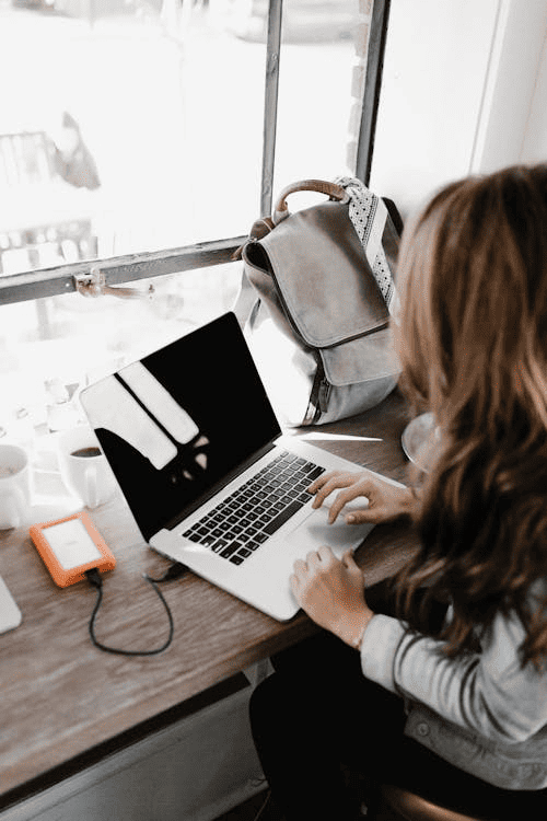 A woman using a laptop while sitting at her work desk near a window. 