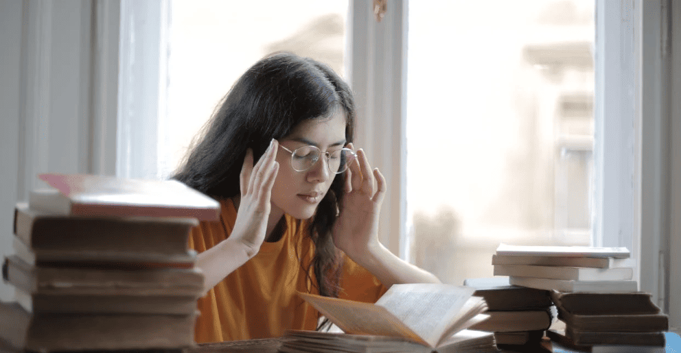 A student sitting in front of books with her hands on her temples.