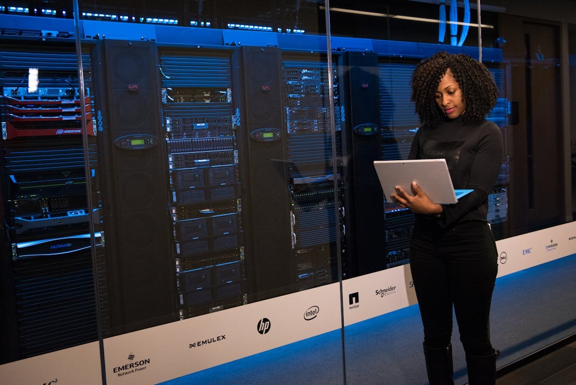  A woman looking at ther laptop while standing next to server units.