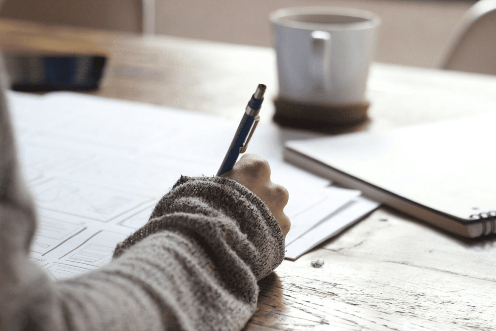 A person writing notes on a piece of paper on top of a brown wooden table near a white ceramic mug.