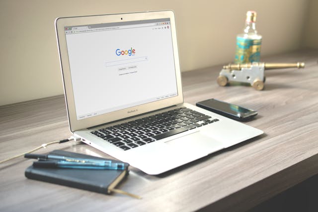 A laptop with Google pulled up in the browser on a desk next to a notebook, phone and trinkets. 