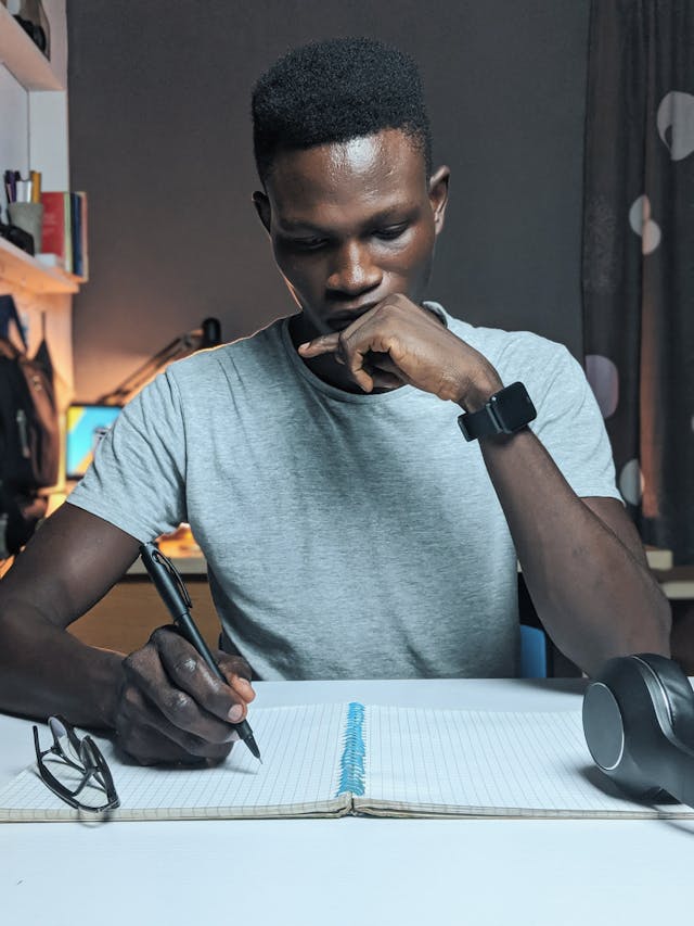 Man at a desk with one hand on his chin, the other hand writing in a notebook as he thinks of what to write in his essay.