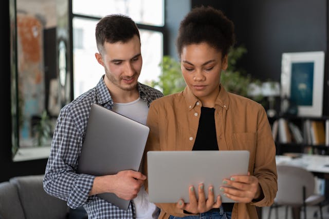 A man holding a laptop looking over the shoulder of a woman holding an open laptop.