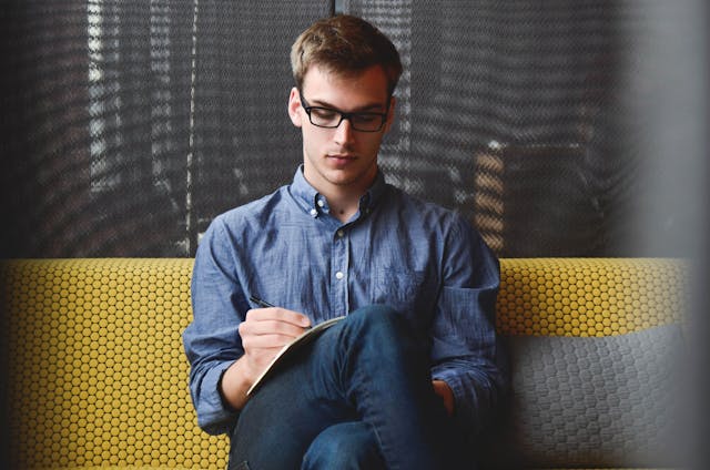 A man wearing glasses sitting on a yellow couch while writing an essay in a notebook.