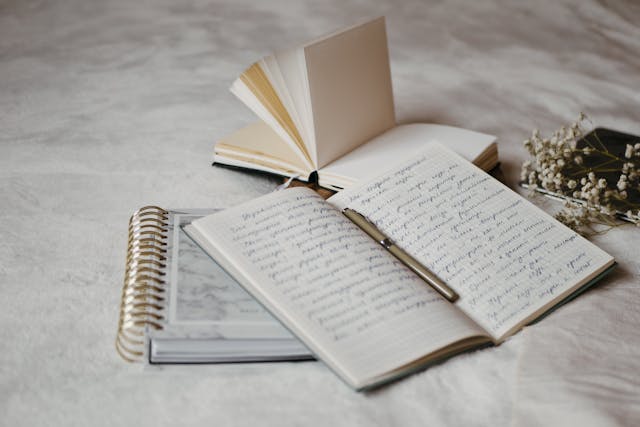 An open notebook with a pen sitting on top of two other notebooks on a textured surface.