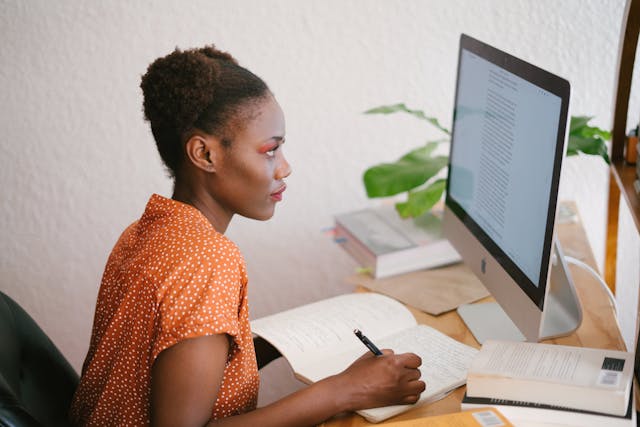 Woman sitting at a desk, looking at a computer screen while writing in a notebook.