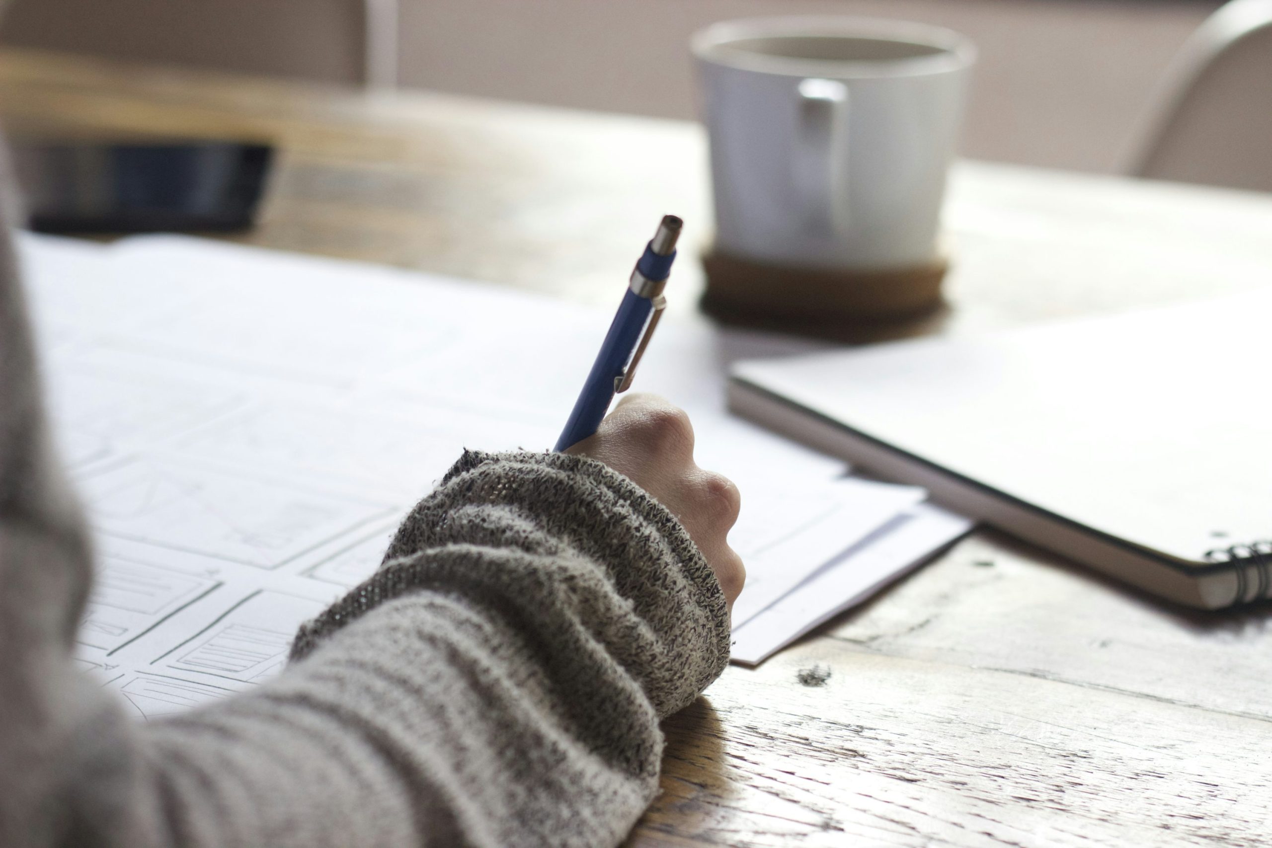 A person writing on paper on a brown wooden table near a white ceramic mug.