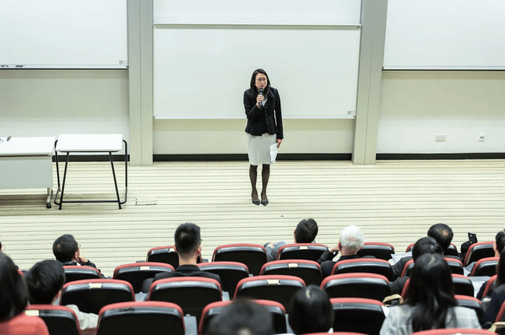 Seorang profesor berdiri di depan kelas mahasiswa. 