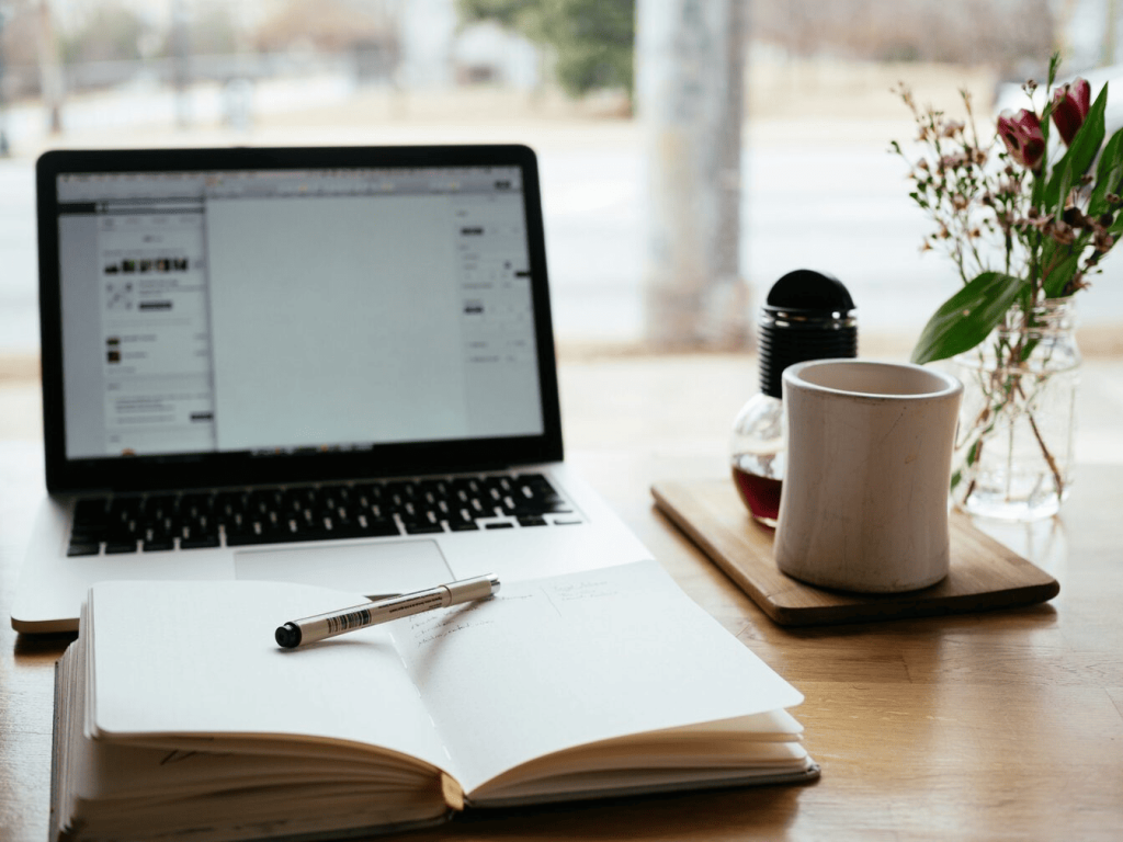 A laptop next to a coffee mug, notebook and pen.