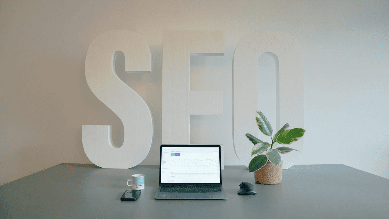 A laptop computer sitting on top of a desk between a plant and coffee cup with the word "SEO" in the background.