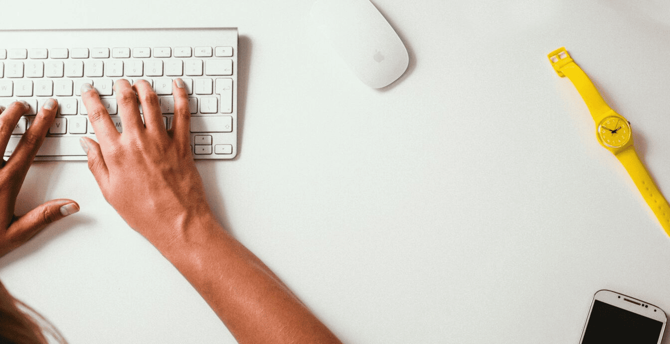 A person typing on an Apple cordless keyboard with a mouse, yellow watch, and smarthone next to them.