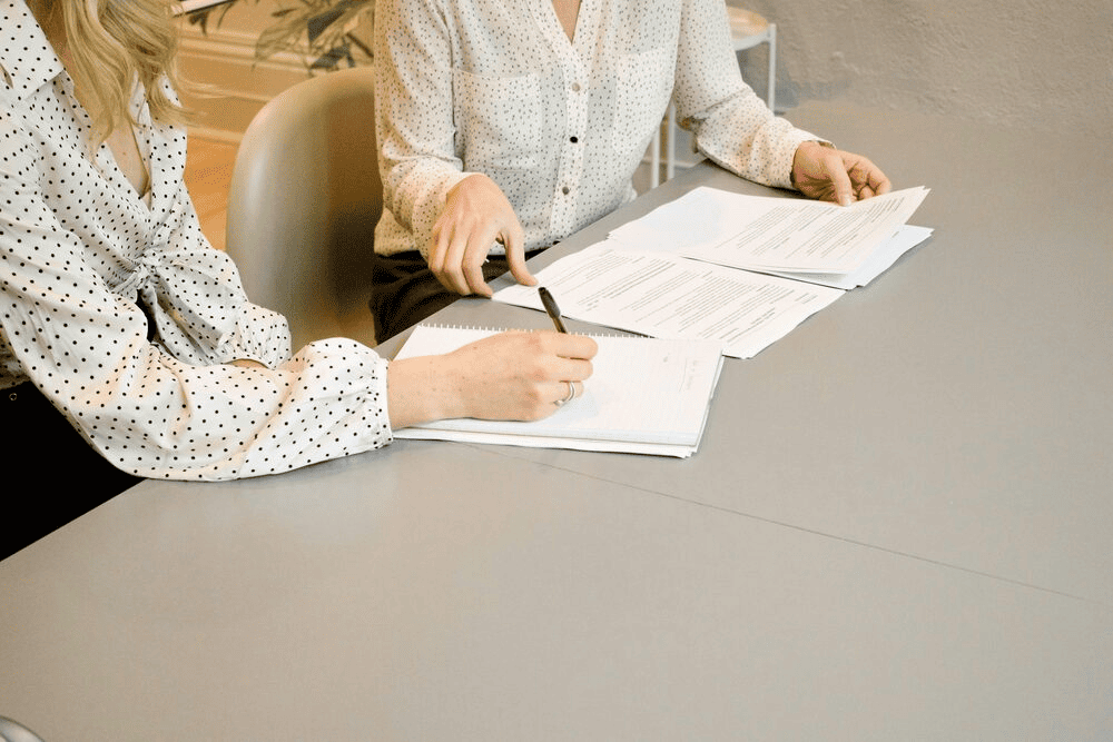 Two women going through some documents, with one writing some notes on paper.