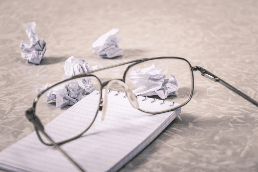 Reading glasses on top of a notebook and surrounded by crampled pieces of paper.