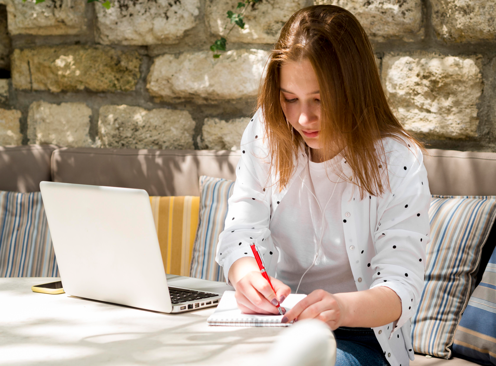一位年轻女性一边使用笔记本电脑,一边在笔记本上写字。